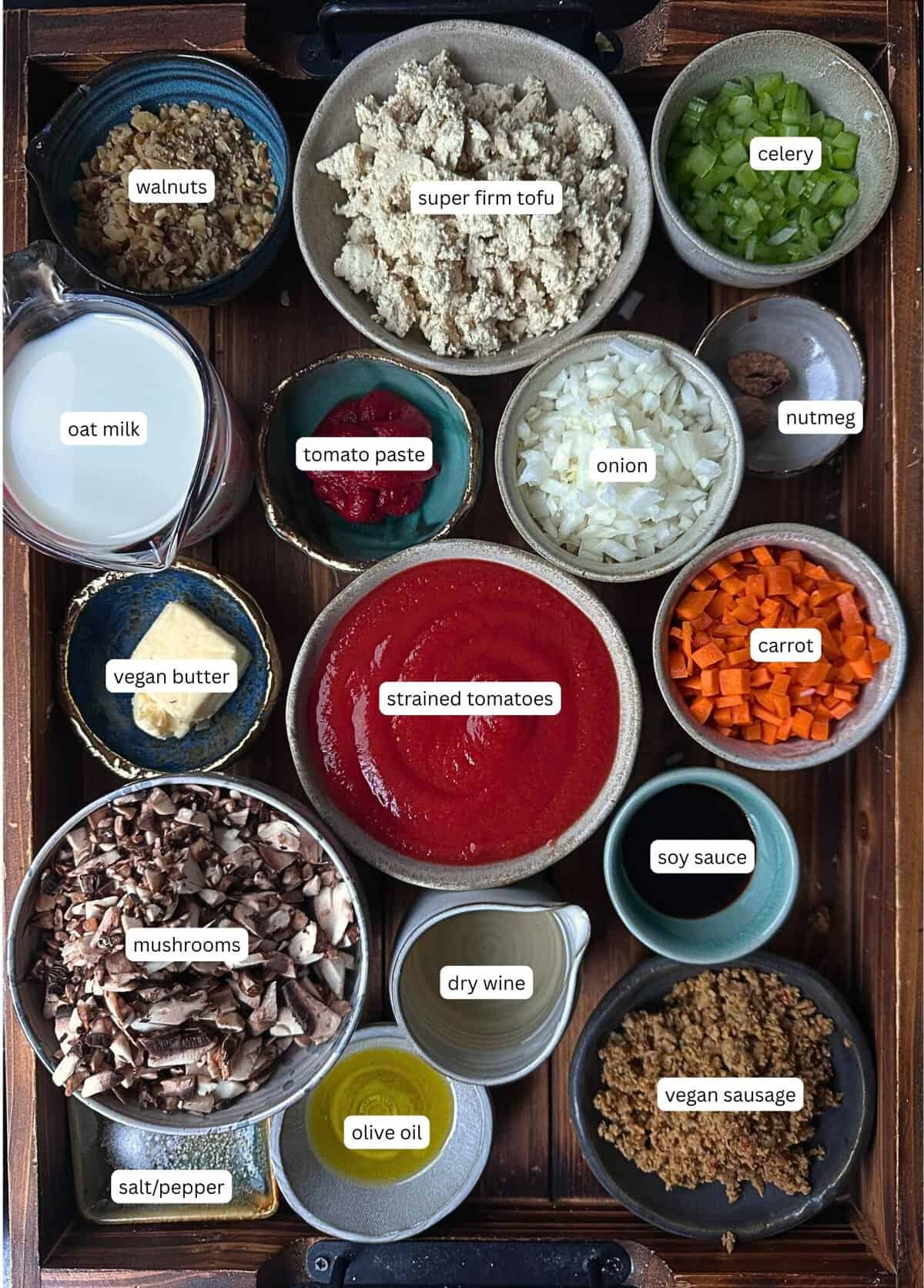 BEST Vegan Bolognese Recipe - Meaty and Delicious. 15 A tray with labeled bowls of ingredients for vegan bolognese, including walnuts, super firm tofu, celery, oat milk, vegan butter, strained tomatoes, tomato paste, onion, nutmeg, carrot, mushrooms, soy sauce, dry wine, olive oil, vegan sausage, and salt/pepper.