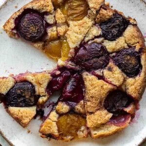 A rustic vegan plum torte with golden brown crust, topped with halved red and yellow plums, sits on a white plate with a slice missing.