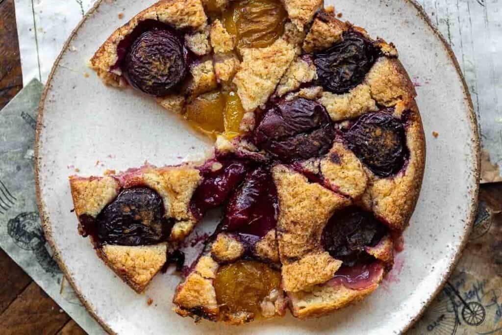 A rustic vegan plum torte with golden brown crust, topped with halved red and yellow plums, sits on a white plate with a slice missing.