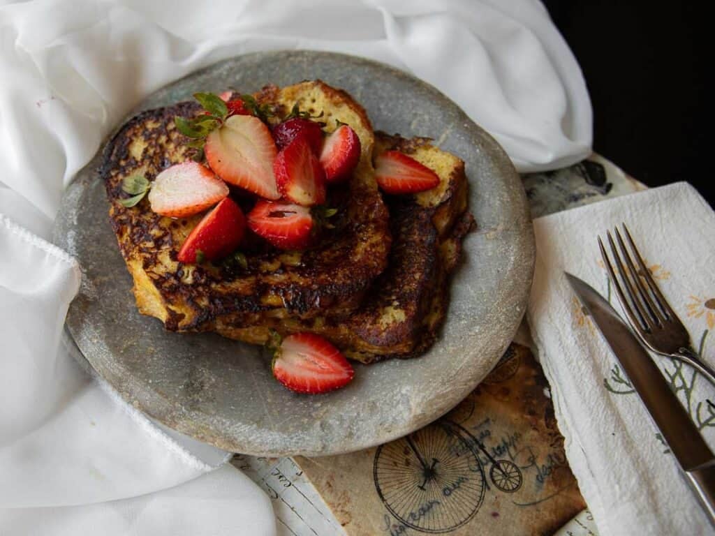 5-Ingredient Vegan French Toast Recipe - Fast & Easy! 14 Two slices of vegan French toast topped with fresh strawberries are served on a rustic gray plate, next to a white napkin with a fork and knife, on a textured surface with white fabric in the background.