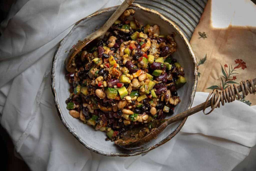 A bowl of colorful Korean BBQ dense bean salad with kidney beans, white beans, black beans, diced vegetables, and herbs, mixed and served with wooden salad utensils on a rustic plate.