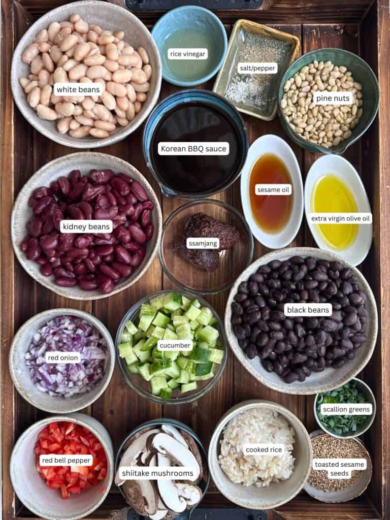 A wooden tray with bowls containing ingredients for dense bean salad: white beans, kidney beans, black beans, red onion, red bell pepper, cucumber, shiitake mushrooms, cooked rice, pine nuts, rice vinegar, salt/pepper, BBQ sauce, ssamjang, oils, scallion greens, and sesame seeds.