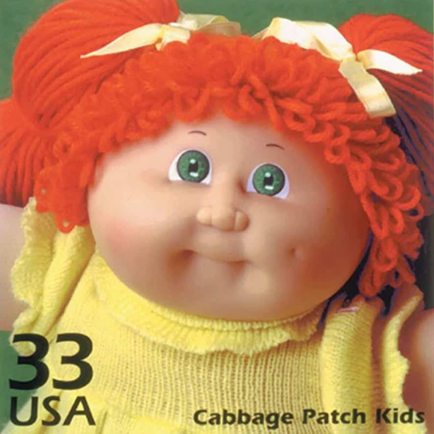 A close-up of a Cabbage Patch Kids doll with green eyes, red yarn hair in pigtails tied with yellow ribbons, wearing a yellow outfit. Text reads: 33 USA Cabbage Patch Kids—just like the one my little brother got from the ice cream truck.