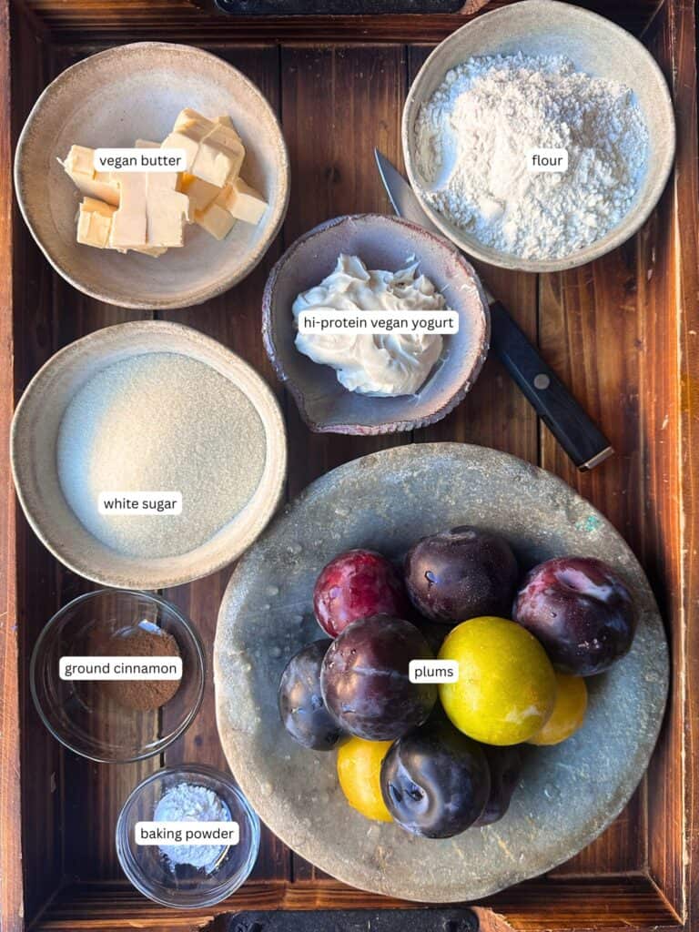 A wooden tray with bowls containing labeled ingredients: vegan butter, flour, white sugar, high-protein vegan yogurt, ground cinnamon, baking powder, and a plate of assorted plums, with a knife beside the yogurt.