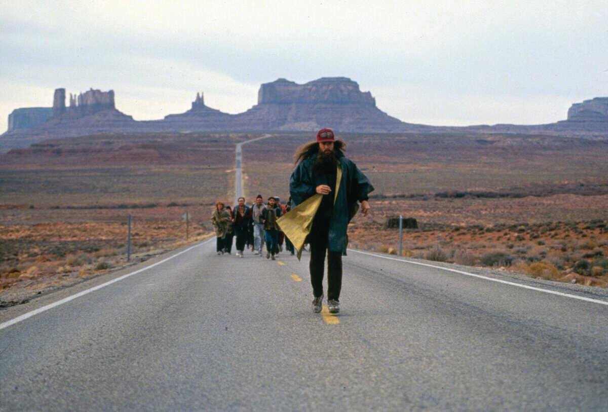 A man with long hair and a red cap runs down an empty desert highway, rock formations towering behind him, as a small group of runners follows his lead.