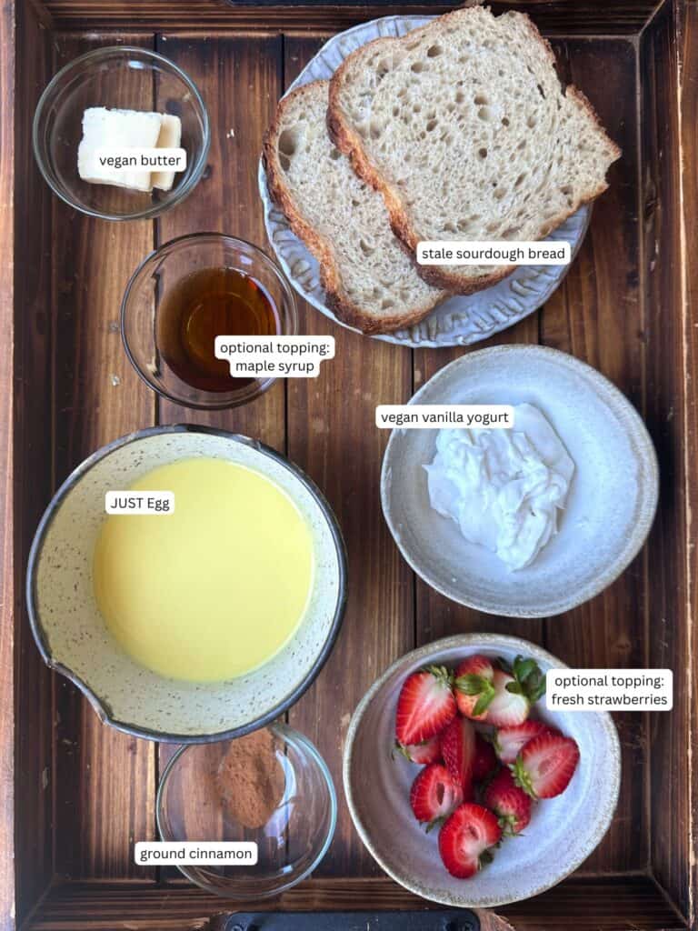 5-Ingredient Vegan French Toast Recipe - Fast & Easy! 16 A wooden tray holds bowls and plates with ingredients for vegan French toast: vegan butter, maple syrup, stale sourdough bread, vegan vanilla yogurt, JUST Egg, strawberries, and ground cinnamon, each labeled.