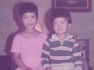 A young Joanne in a pink dress and her little brother Jaesun, in a striped shirt and light pants, sit side by side on a wooden bench, smiling in a warmly lit room with patterned wallpaper and a framed picture in the background.