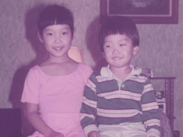 A young Joanne in a pink dress and her little brother Jaesun, in a striped shirt and light pants, sit side by side on a wooden bench, smiling in a warmly lit room with patterned wallpaper and a framed picture in the background.