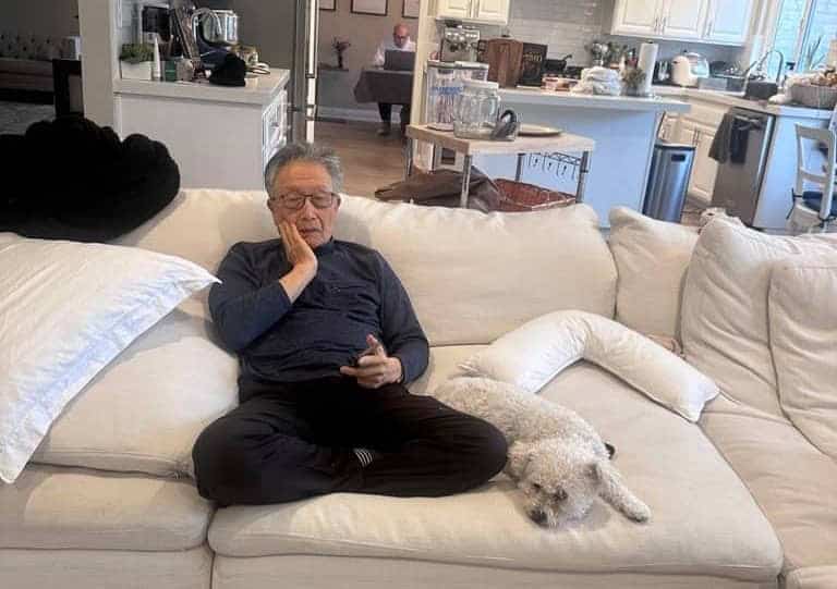 An older man sits cross-legged on a white couch, holding his face while looking at his phone, perhaps scrolling through social media. A small white dog lies next to him. In the background, another person works at a table in the kitchen.