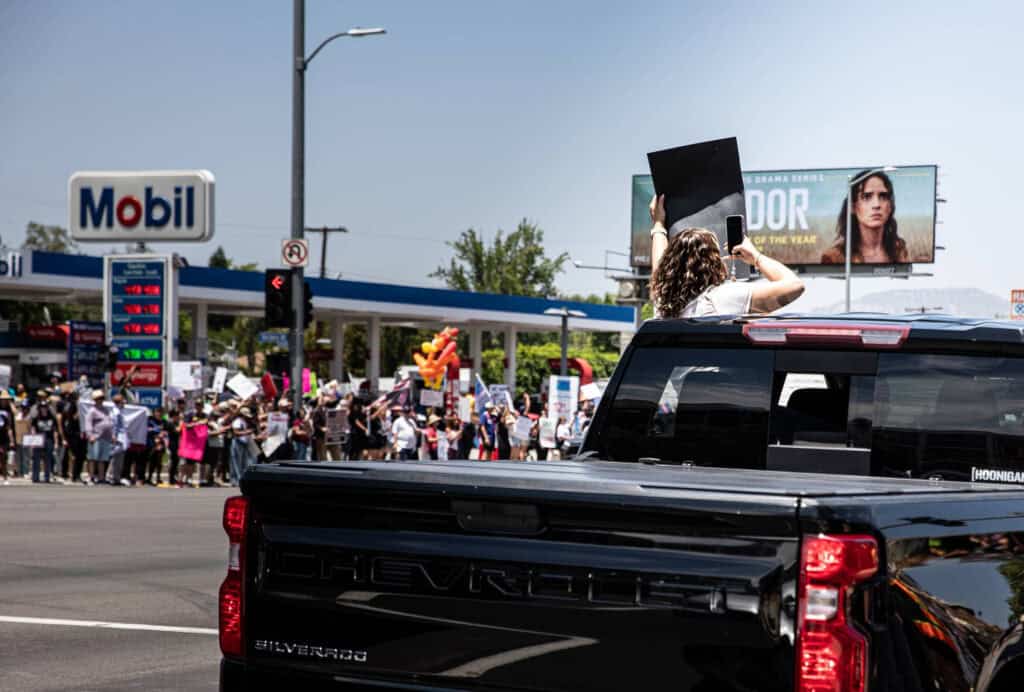 A person stands through the sunroof of a black Chevrolet truck holding a sign at a busy intersection, while protesters with signs, driven by political causes  gather near a Mobil gas station in the background.