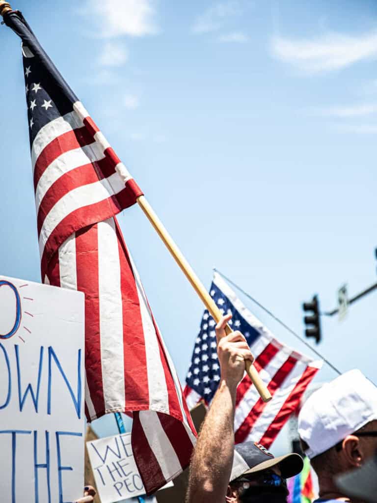 A person holds up a large American flag at a political rally, surrounded by others with signs and flags, as the vibrant scene is captured against a backdrop of a blue sky and traffic light.