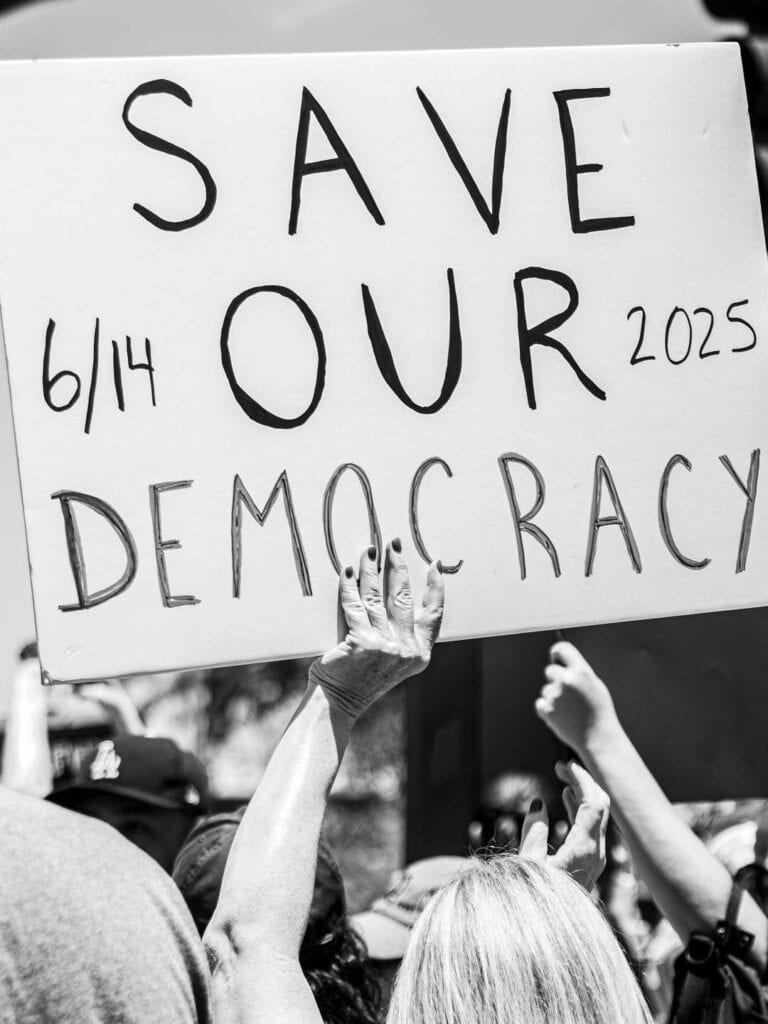 A black-and-white photo of people at a political rally, with a person holding a sign that reads SAVE OUR DEMOCRACY 6/14 2025, surrounded by raised hands—capturing the power of collective action in the age of social media.