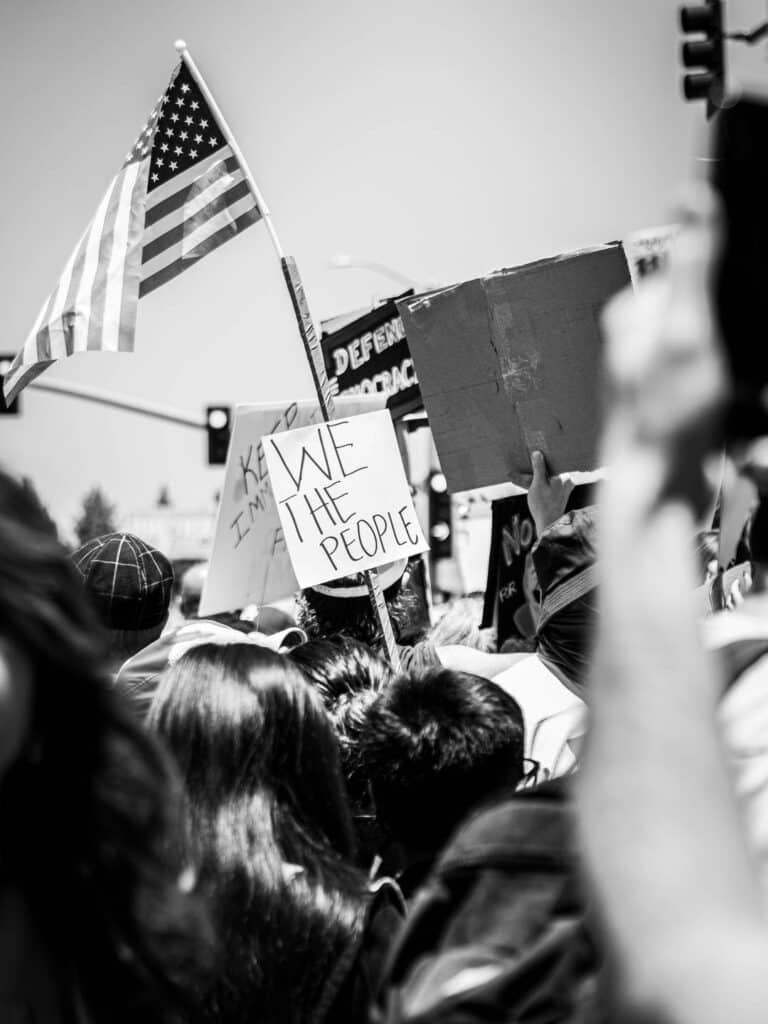 A black-and-white photo of a protest crowd holding signs, including one that reads WE THE PEOPLE and another with an American flag. The energetic, united scene highlights the power of social media in driving political movements under bright daylight.