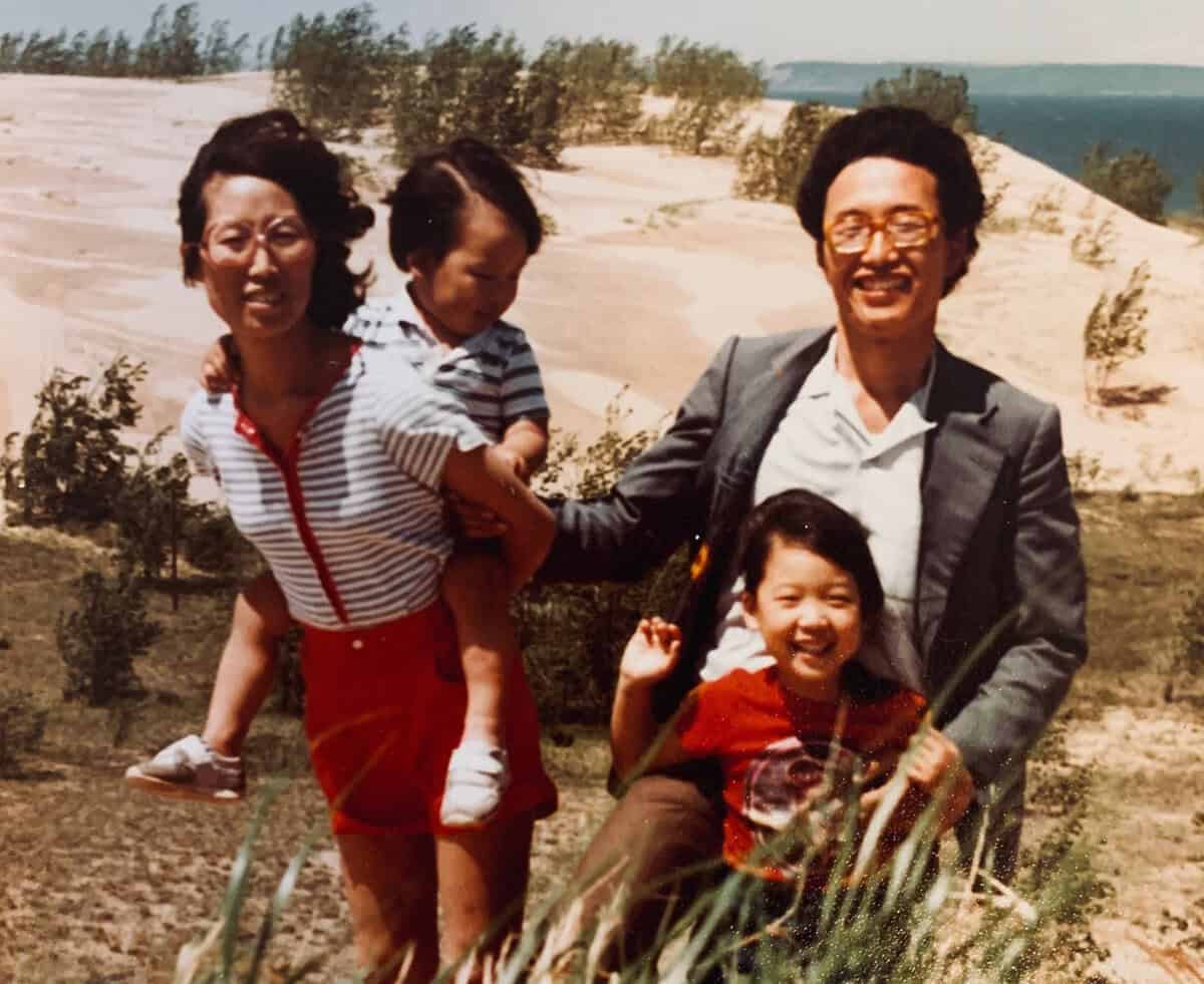A smiling family of four poses outdoors near sand dunes and greenery. The mother holds a young child, while the father, in a suit, holds another child who is smiling and flashing a peace sign. The sky is clear and blue.