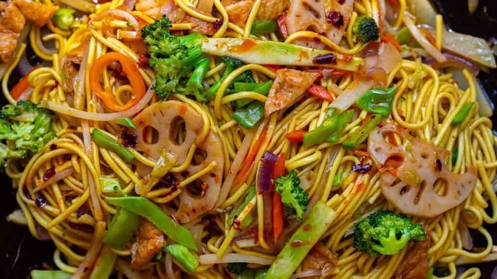 Easy & Healthy Lo Mein Vegan Recipe: Done in Minutes. 17 A close-up of colorful stir-fried lo mein vegan noodles mixed with broccoli, carrots, lotus root slices, snow peas, and pieces of tofu, all garnished with chili flakes.
