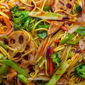 Easy & Healthy Lo Mein Vegan Recipe: Done in Minutes. 37 Close-up of stir-fried lo mein vegan noodles with broccoli, sliced lotus root, red onion, carrots, snow peas, and pieces of tofu, coated in a savory sauce with visible chili flakes.