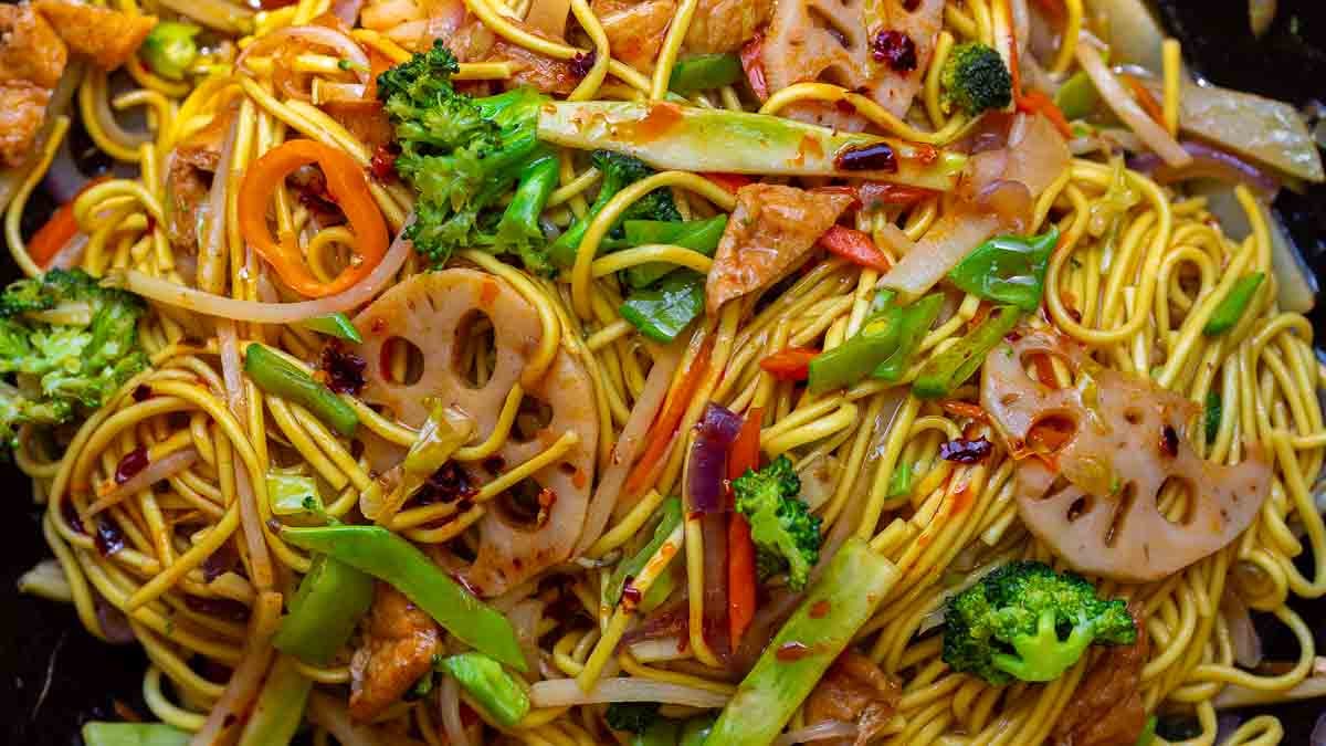 A close-up of colorful stir-fried lo mein vegan noodles mixed with broccoli, carrots, lotus root slices, snow peas, and pieces of tofu, all garnished with chili flakes.
