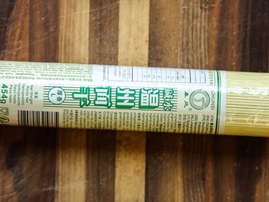 Easy & Healthy Lo Mein Vegan Recipe: Done in Minutes. 20 A package of dried noodles with green Chinese text and a nutrition label, lying on a wooden surface. The clear packaging reveals the yellow noodles inside.