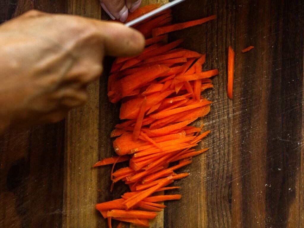 A hand slices a carrot into thin strips on a wooden cutting board with a kitchen knife.