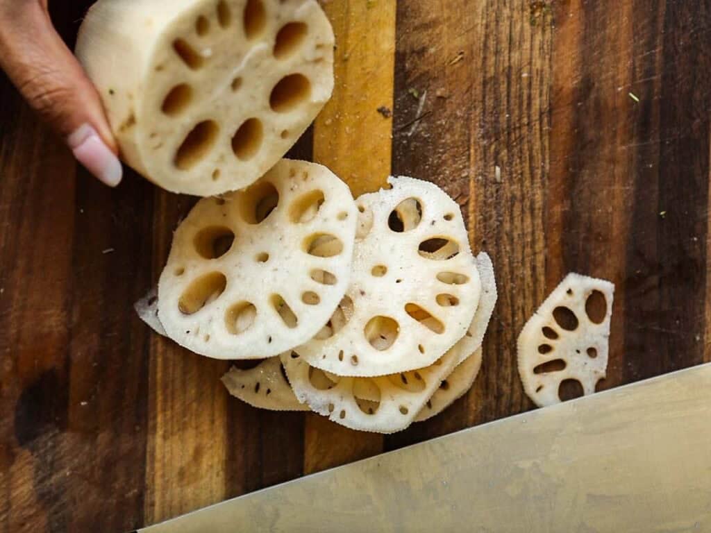 A hand holds a piece of lotus root above a wooden cutting board, with several round slices of lotus root showing their distinctive holes. A knife is visible at the bottom right.