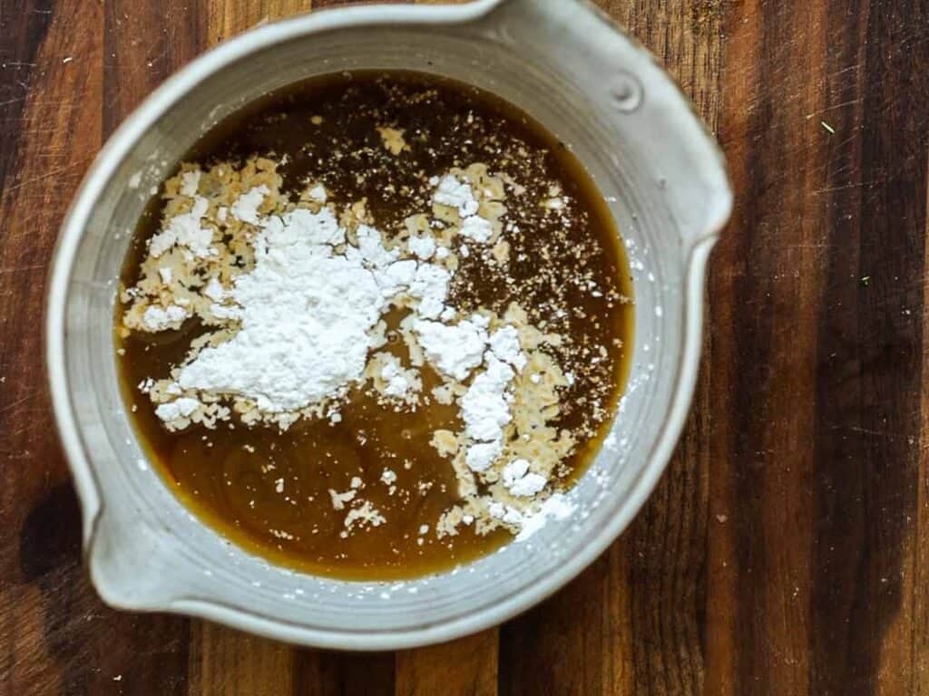 A ceramic mixing bowl with a brown liquid and corn starch on top sits on a wooden surface, ready to be blended.