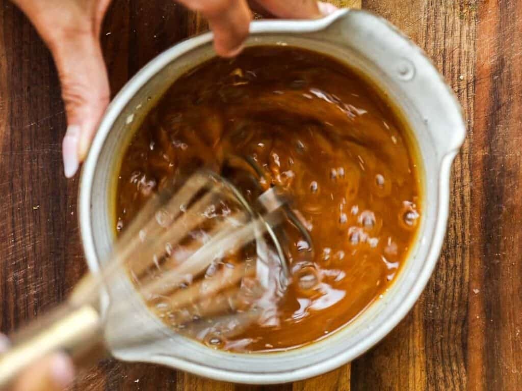 A person uses a whisk to stir a thick, brown sauce or mixture in a white bowl on a wooden surface.