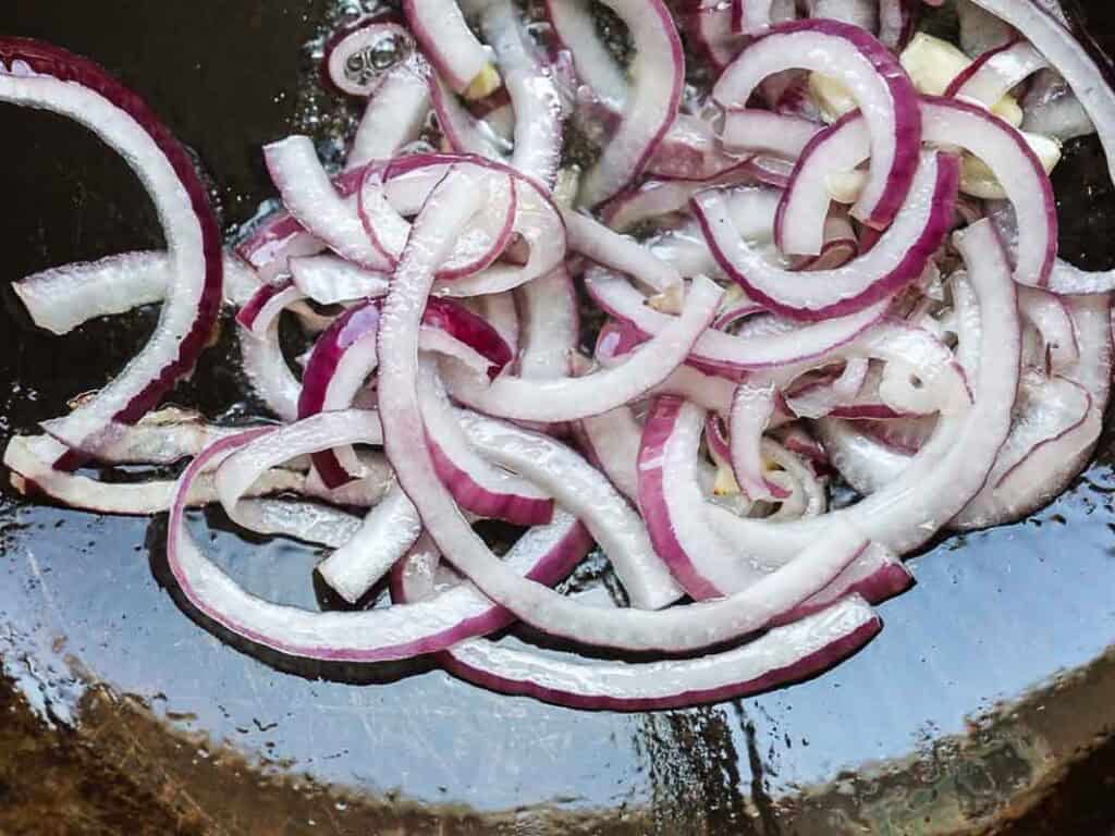 Sliced red onions cooking in a pan, starting to soften and turn translucent with some oil glistening around them.