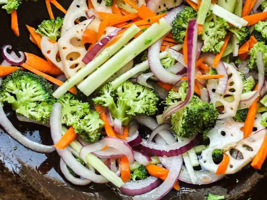 A colorful vegetable stir-fry with broccoli, red onion, carrot sticks, and sliced lotus root in a dark pan.