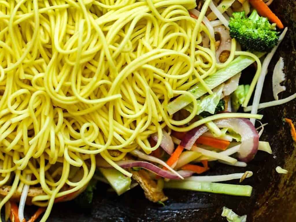 Close-up of yellow noodles resting on top of mixed stir-fried vegetables, including sliced red onions, carrots, broccoli, and cabbage in a dark pan.