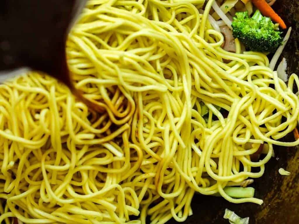 Close-up of yellow noodles being topped with a dark sauce, with visible pieces of broccoli, carrots, and bean sprouts mixed in.