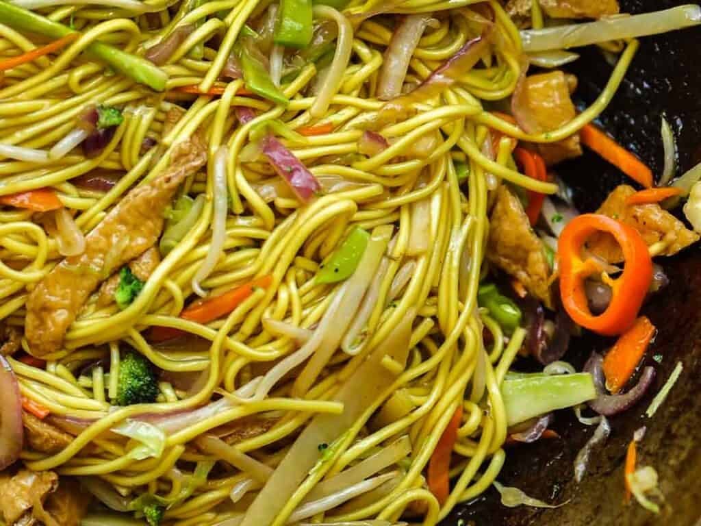 Easy & Healthy Lo Mein Vegan Recipe: Done in Minutes. 19 Close-up of vegetable lo mein vegan noodles mixed with sliced red peppers, carrots, broccoli, onions, bean sprouts, and tofu in a wok. The colorful vegetables are intertwined with the yellow noodles.