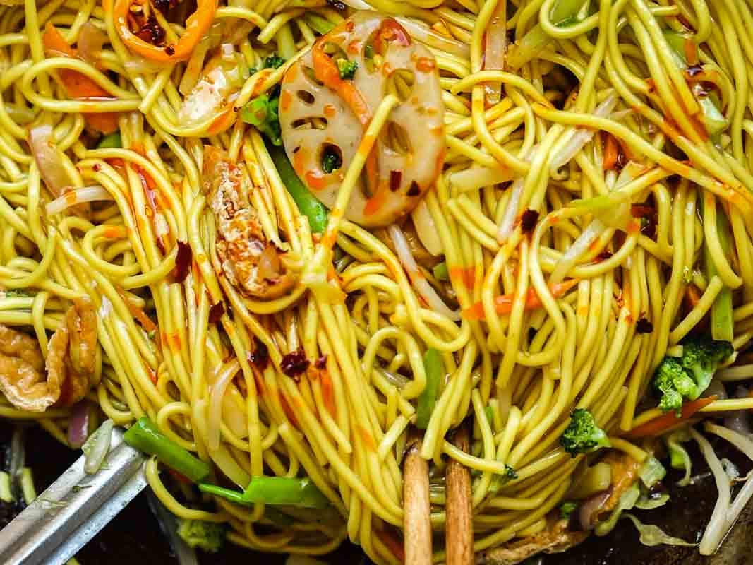 Easy & Healthy Lo Mein Vegan Recipe: Done in Minutes. 26 A close-up of stir-fried lo mein vegan noodles mixed with sliced lotus root, broccoli, carrots, onions, and other vegetables, coated in a savory sauce.