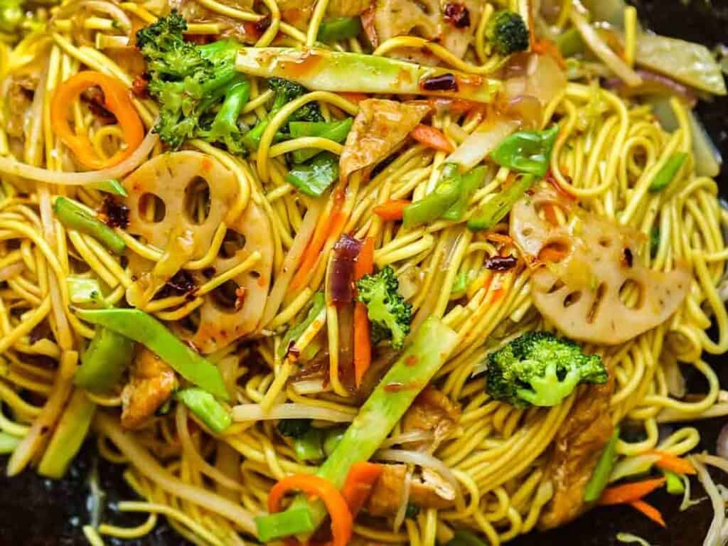 Easy & Healthy Lo Mein Vegan Recipe: Done in Minutes. 18 A close-up of stir-fried lo mein vegan noodles mixed with sliced lotus root, broccoli, carrots, onions, and other vegetables, coated in a savory sauce.