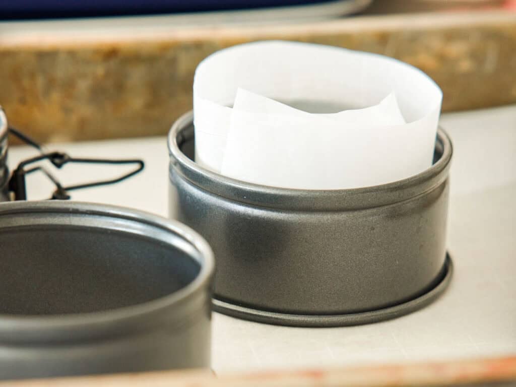 A round metal baking pan with a strip of white parchment paper lining the inside, placed on a light surface with another similar pan nearby.