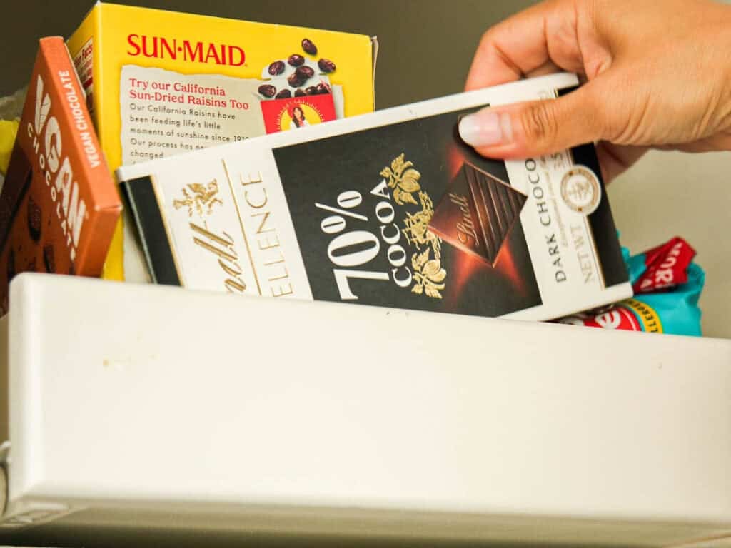 A hand reaches into a drawer filled with chocolate bars and a box of Sun-Maid raisins, selecting a Lindt 70% cocoa dark chocolate bar.
