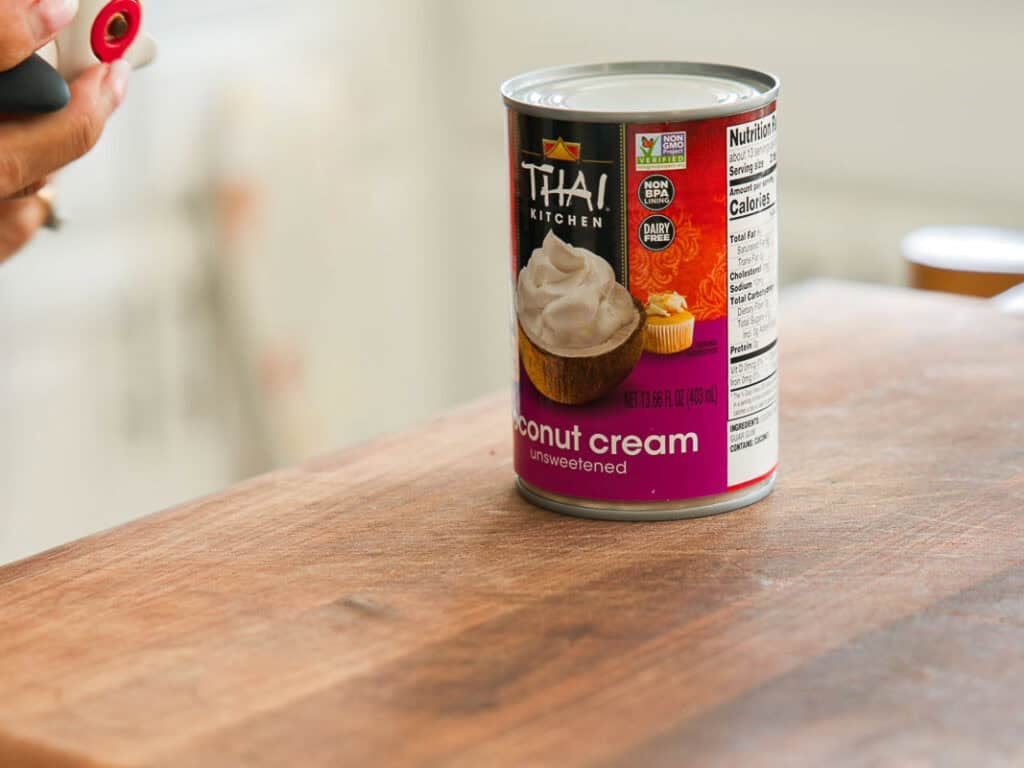 A can of Thai Kitchen unsweetened coconut cream sits on a wooden countertop, with a person holding a can opener nearby. The label shows cupcakes topped with whipped cream.