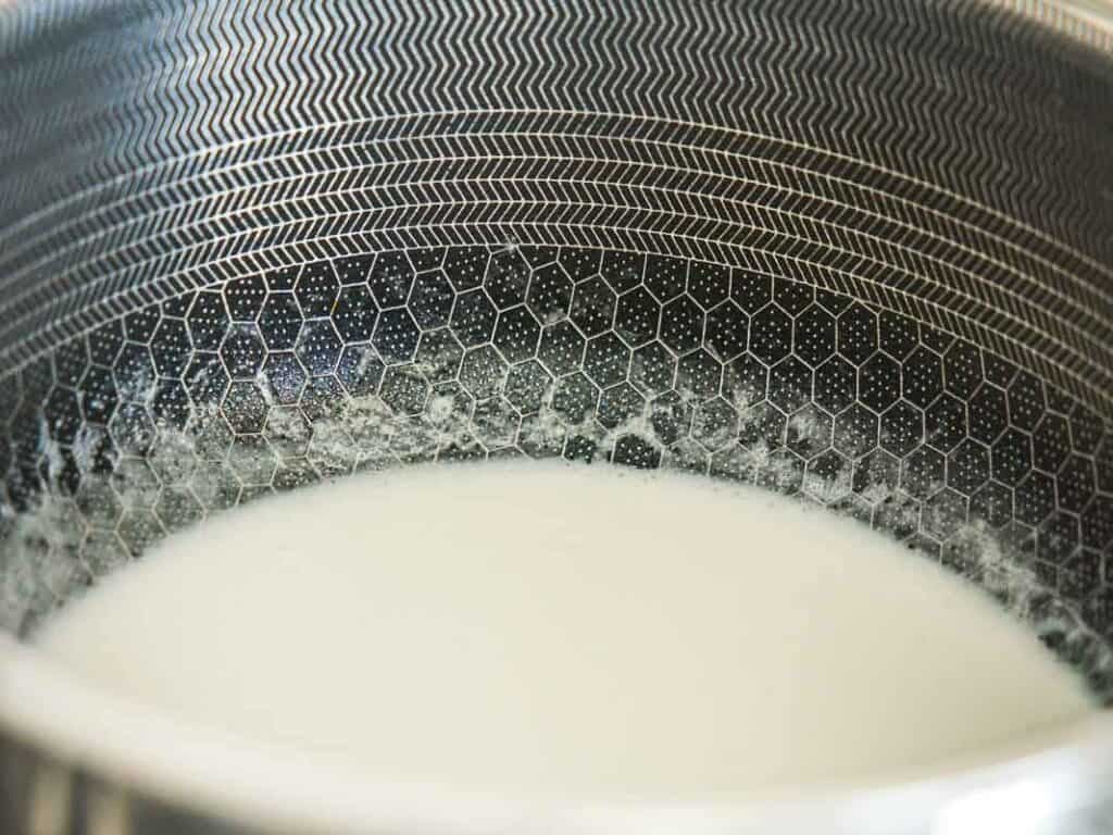 Close-up of coconut cream heating in a stainless steel pot with a hexagonal pattern on the inner surface. White milk froth forms along the edge of the pot.