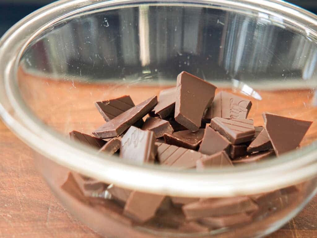 A clear glass bowl filled with broken pieces of dark chocolate sits on a wooden surface.