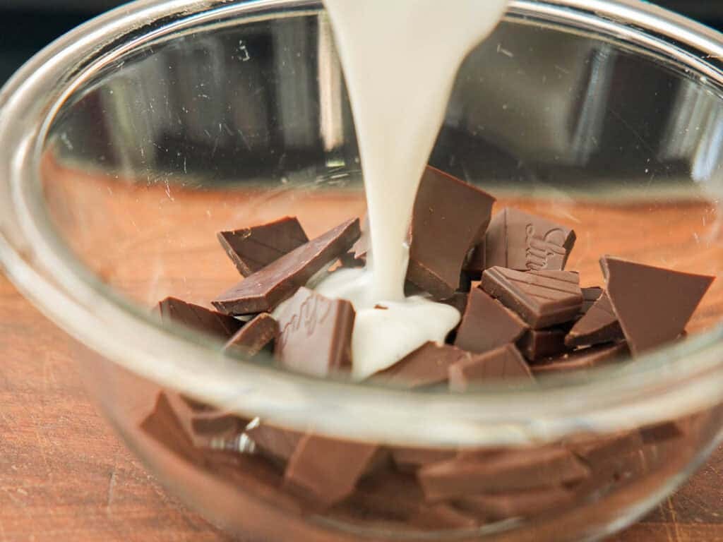 A glass bowl filled with chunks of chocolate as a stream of cream is being poured over them, set on a wooden surface.