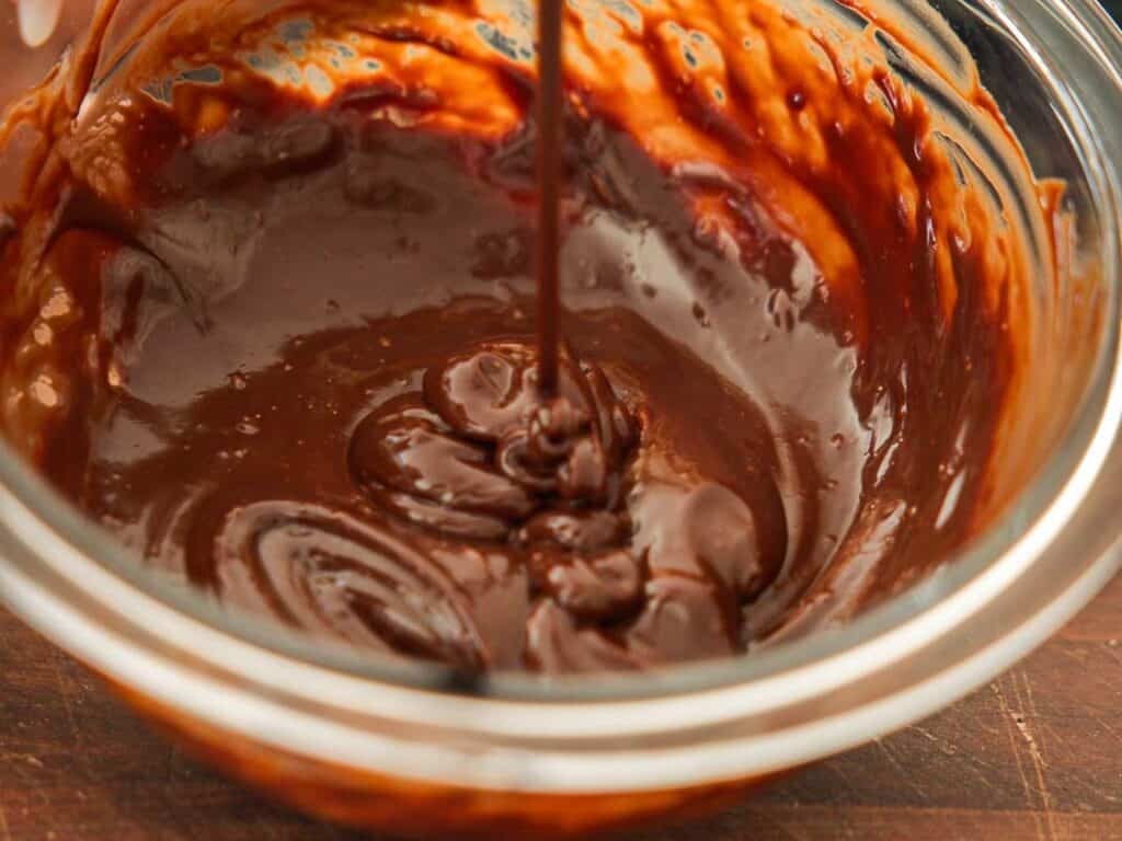 A close-up of thick, glossy melted chocolate being stirred in a glass bowl, with some chocolate being poured in from above. The bowl sits on a wooden surface.