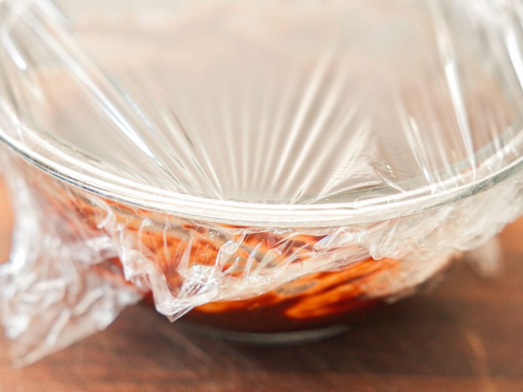 A glass bowl covered with clear plastic wrap containing melted chocolate ganache, placed on a wooden surface.