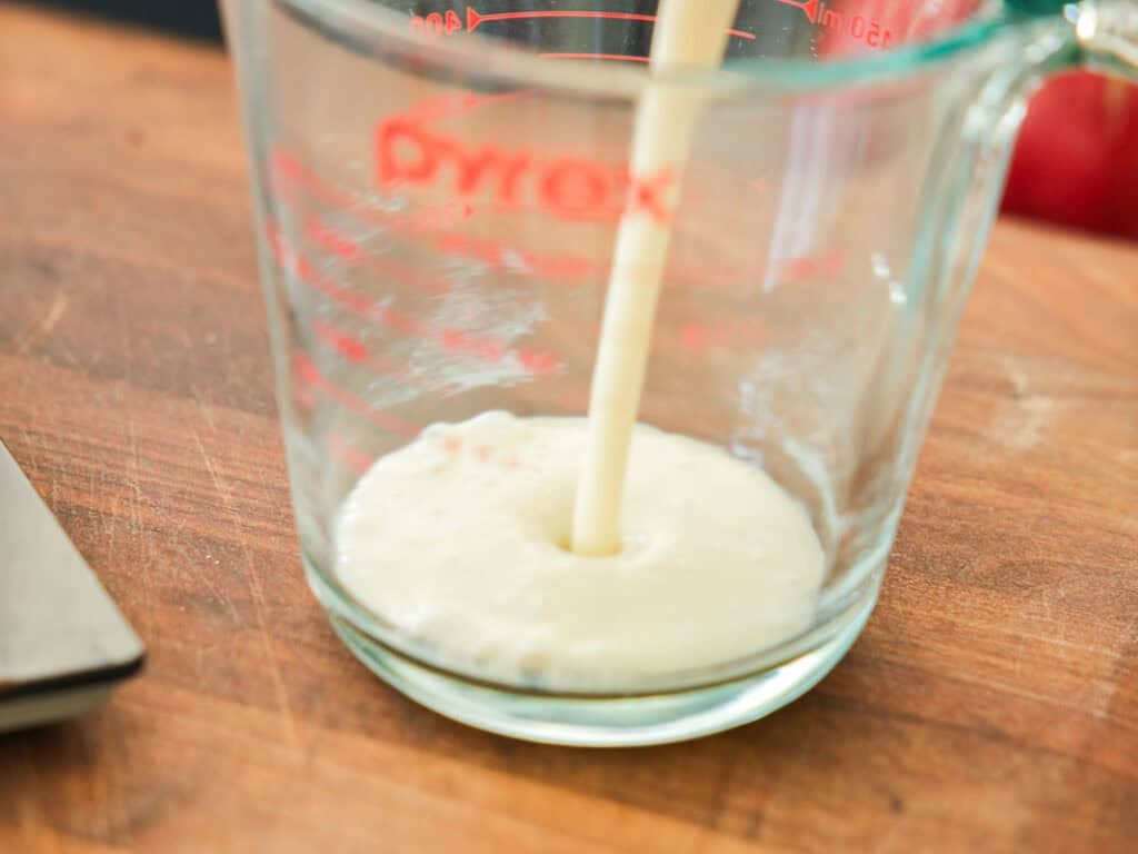 Soy milk being poured into a clear glass Pyrex measuring cup, sitting on a wooden surface.