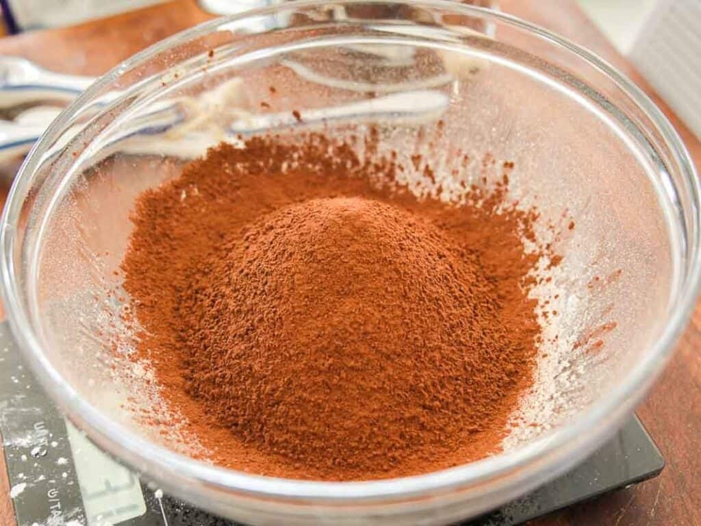 A glass mixing bowl filled with a mound of sifted cocoa powder sits on a kitchen counter, ready for baking.