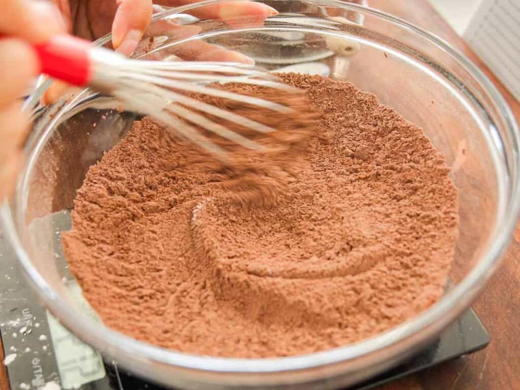 A hand using a whisk to mix cocoa powder and flour in a clear glass bowl on a kitchen counter.