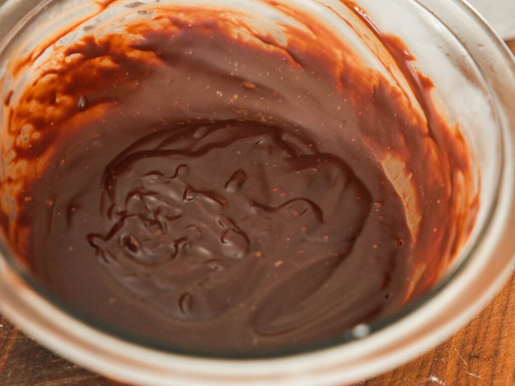 A glass bowl filled with smooth, glossy hardened chocolate ganache, with some streaks and swirls visible along the edges. The bowl is resting on a wooden surface.