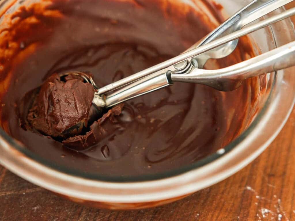 A metal scoop holds a ball of thick chocolate mixture over a glass bowl filled with melted chocolate on a wooden surface.