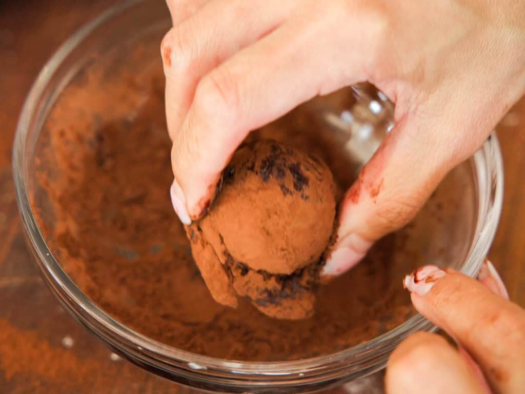 A close-up of hands rolling a chocolate truffle in cocoa powder inside a glass bowl. The truffle and fingers are lightly dusted with cocoa.