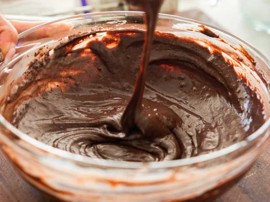 A close-up of thick chocolate cake batter being mixed in a clear glass bowl, with some batter being lifted by a spoon or spatula.