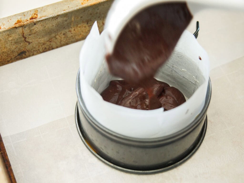 A close-up of chocolate cake batter being poured from a spoon into a round, parchment-lined baking pan on a baking sheet.