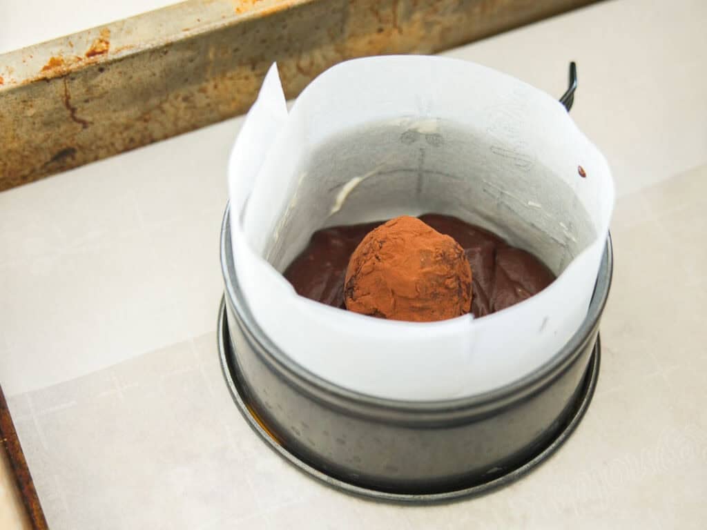 A chocolate truffle coated in cocoa powder sits on top of chocolate batter in a parchment-lined round baking pan on a metal tray.